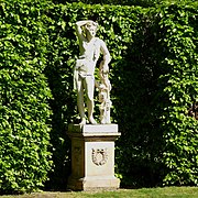 statue on a plinth in front of a green hedge. A youth in classical dress leans against a tree trunk