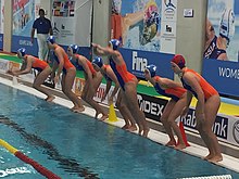 Sieben Niederländerinnen im orangefarbenen Badeanzug springen vom Beckenrand ins Wasser. Die Torhüterin trägt eine rote Bademütze, die Feldspielerinnen tragen eine blaue Kappe.