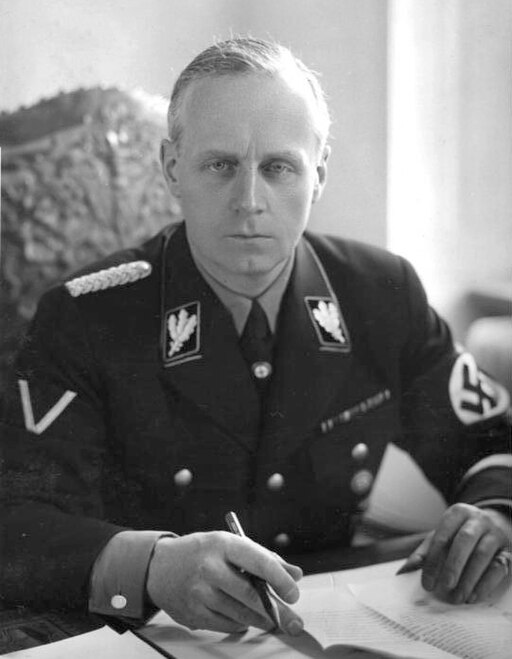 Portrait of the middle-aged man with short grey hair and a stern expression. He wears a dark military uniform, with a swastika on one arm. He is seated with his hands on a table with several papers on it, holding a pen.