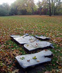 5 unregelmäßig geformte, mehrere Meter große Bleiplatten liegen dicht beieinander auf einer herbstlichen Wiese in einem Stadtpark.