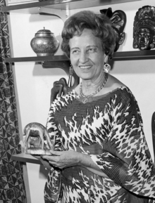 a woman dressed in African dress and jewelry holds a small sculpture of an African animal. Other artifacts line the shelves behind her.