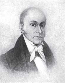 A man, bald on top with dark hair in the back, wearing a dark jacket and white shirt