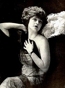 A young white woman with curly bobbed hair, one hand behind her neck, one on her chest