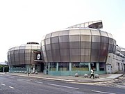 View of two of the four stainless steel drums from across an empty road. The main entrance is visible between the two drums with a circular sign above containing the words "the HUBS".