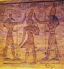 Relief of a man with an elaborate crown between two figures who gesture toward the crown. The figure on the left has the head of an animal with square ears and a long nose, while the one on the right has a falcon's head.