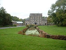 canal-type waterway running through a meadow to a large grey four-storey building in the distance