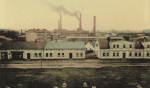 Archiwalne zdjęcie przedstawia zabudowania fabryki celulozy. Na pierwszym planie znajduje się rząd parterowych, białych budynków, nakrytych wysokimi dachami. Za nimi są wyższe, ceglane zabudowania. Nad całą fabryką góruje pięć wysokich kominów. Z trzech z nich unosi się gęsty dym.