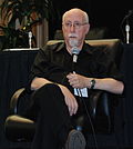 White middle-aged man sitting on black seat with leg crossed, holding a microphone with left hand, while the right hand rests on his legs.