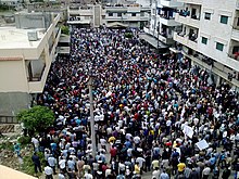 A street between medium-height modern white apartment blocks filled with people, some carrying signs