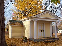 A one-story yellow wooden building with a pointed roof held up by columns at the front and a small addition on the left in a cemetery. In the front is a wooden door with "1823" on it. It is autumn, and the ground and roof are covered with yellow fallen leaves. A tree with yellow leaves and an American flag are visible in the background