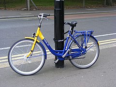 Farbfoto. Blau-gelbes Hollandrad, tiefer Einstieg, Beleuchtung, Gepäckträger, Handbremse, keine Gangschaltung. Auf dem Rahmen steht „OV-fiets.nl“. Angeschlossen an Laternenpfahl mit fest am Sattel montiertem Kettenschloss. Im Hintergrund eine Straße.