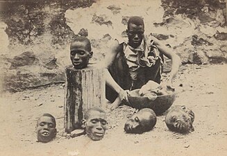 Photographie présentant un homme assis devant quatre têtes humaines coupées, au sol, et une cinquième sur un billot de bois dressé.