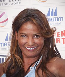 A woman with brown hair wearing a blue bikini top. She is smiling wide.