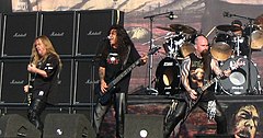 The band Slayer is shown at concert. From left to right are an electric guitarist, a bass player (also singing), an electric guitarists, and a drummer. The first guitarist and bassist have long hair. The right-most guitarist has a bald head. The drummer has two bass drums.