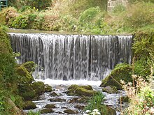 A weir with a river cutting a rough winding channel roughly 2 metres deep flowing through a wooded valley with paths.