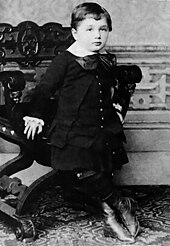 Un muchacho joven con el pelo corto y una cara redonda, con un arco de cuello blanco y grande, con chaleco, chaqueta, falda y botas altas.  Está apoyado en una silla ornamentada.