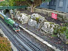 A green miniature train engine pulls carriages under a bridge and past a rocky hill