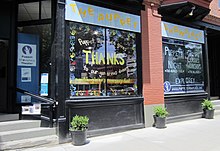 The exterior and sidewalk of a brick building. Above two large windows hangs a sign with the words "THE PUPPET SHOWPLACE." The left window is painted with the words, "Puppet Showplace gives THANKS to our generous donors." The right window is painted with the words, "PUPPETS AT NIGHT. CAMPS. CLASSES. WORKSHOPS." Steps lead up to a glass door through which the Puppet Showplace Theater logo is visible.