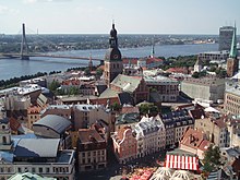 Farbfotografie in der Obersicht von einer Stadt mit vielen alten Häusern und engen Gassen im Vordergrund. In der Mitte ist eine braune Kirche mit dunkler Spitze. Im Hintergrund ist ein Fluss mit einer Brücke an der linken Seite zu sehen, die zum anderen Ufer mit Gebäuden und Bäumen führt.