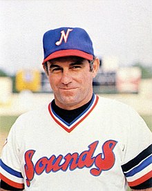 A man in a blue cap with a white "N" on the front and a white baseball jersey with "Sounds" across the chest in red and blue smiles.