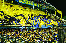 Photographie en couleurs. Dans les tribunes d'un stade, de nombreux supporters agitent des banderoles de couleur jaune et bleue.