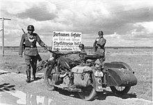 Trois hommes en uniforme, à moto, devant un panneau.