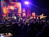 A group of seven musicians are sitting on a stage, performing with acoustic instruments