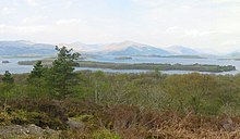 Low trees and bushes in the foreground give way to a body of water with a large wooded isle close to the shore and smaller islets beyond. There are mountains in the distance.