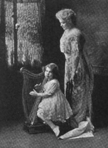 A white woman with light hair in a bouffant style, wearing a lacy light-colored dress, photographed in profile against a dark background; she is gazing downward at a small white girl playing a scaled-down harp