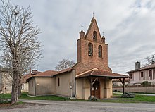 Photo de l'église Saint-Loube. Vue de la porte d'entrée, protégée par un porche et située sur le clocher-mur en briques qui porte trois cloches et trois croix. L'édifice est une église à nef unique avec un toit en tuiles.