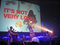 Рок-группа выступает на сцене с надписью «ЭТО НЕ ОЧЕНЬ ГРОМКО» на экране позади них.