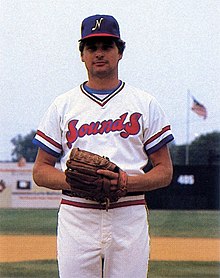 A man in a white baseball uniform with "Sounds" on the front in red and blue and a blue cap with a white "N" on the front standing on a field with his hands together in his glove