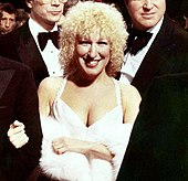 A woman with blonde curly hair wearing a white dress and fur shawl, surrounded by men in tuxedos