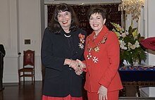 Two smiling women face the camera shaking hands. They are wearing formal clothes.