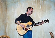 Johnston stands and plays acoustic guitar in the corner of a room.