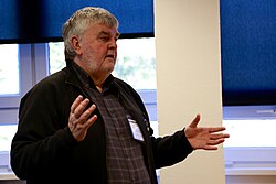 A man with white hair and beard, wearing a checked shirt, a jacket and a name badge speaks whilst standing in a classroom, in front of some windows half covered with blinds on a sunny day.