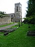 The east end of a stone church is seen through a graveyard from the north. On the right is a battlemented tower and the nave extends to the left