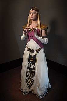 Femme blonde sur un fond noir, vêtue d'une robe blanche et violette et d'une ceinture, c'est un Cosplay de la Princesse Zelda.