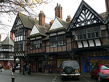 A row of shops seen at an angle with an arcade at ground level. Above this the building is timber-framed, with four gables; the lateral two of these have painted plaster panels and decorated timber framing within the gable, and the central gables contain brick nogging and pargeting.