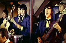 A band consisting of five men wearing royal blue suits, white shirts and black ties, is performing in front of a beige-and-brown wall. Two men (far left, fourth from left) are playing guitars, while two others (middle, far right) are playing percussion instruments. Four band members have dark brown or black hair; the man on the far right is blond. A pole in the foreground separates the three men on the left from the two on the right.