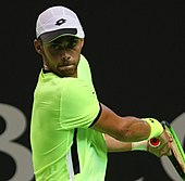 A man in a neon yellow shirt, white shorts, and white hat holds his tennis racket as he prepares to swing
