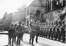 Photographie en noir et blanc d'Adolf Hitler saluant une rangée de soldats en faction devant un bâtiment ancien.