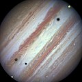 Devant Jupiter, des sphères grises, marron et jaunes sont visibles. Leurs ombres sont projetées sur l'atmosphère de la planète géante.