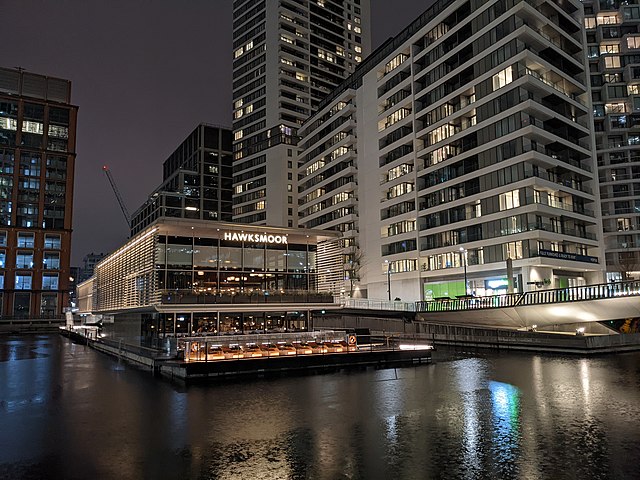 A two storey pavilion floating in a dock, surrounded by tall buildings. On the front is an illuminated sign reading "Hawksmoor" in capital letters
