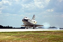 A spaceplane, coloured white on its topside and black on its underside, lands on a runway. A strip of turf is visible in the foreground, there are trees in the background and there is a cloud of smoke coming from the spaceplane's rear wheels.