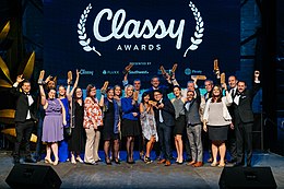 A photograph of twenty people standing in two rows on a stage. They are nicely dressed and holding up award statuettes. The lighting is very blue and behind them is a screen with the Classy logo and sponsors projected.