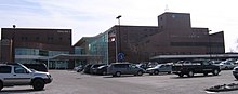 Behind a large parking lot, sits an 8-story tall red brick building with a large number of windows. The words Genesis are on the center of the building at the top. To the left is a slightly shorter building connected to the first with many glass windows. The second building say Pavilion 1