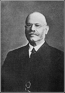 Black and white photograph of man with bald head, sideburns and mustache wearing eyeglasses and a black suit with a white shirt and tie.