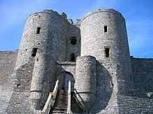 Duas torres de pedra cilíndricas flanqueando uma entrada, e atrás delas duas torres cilíndricas maiores. Um caminho leva ao gateway e as paredes das cortinas estão ligadas às torres.