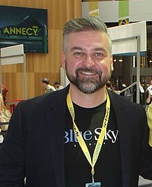 Photo de Troy Quane. Coiffé court, les cheveux et la barbe poivre et sel, la peau claire, il sourit à l'objectif. Il porte un T-shirt arborant le logo de Blue Sky Studio. On devine à l'arrière-plan une salle de conférences.